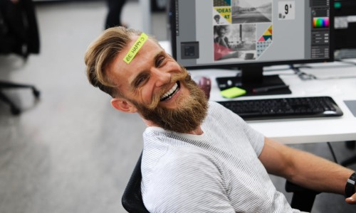 Happy Employee at this desk with a sticky note on his forehead that says "Be Happy"