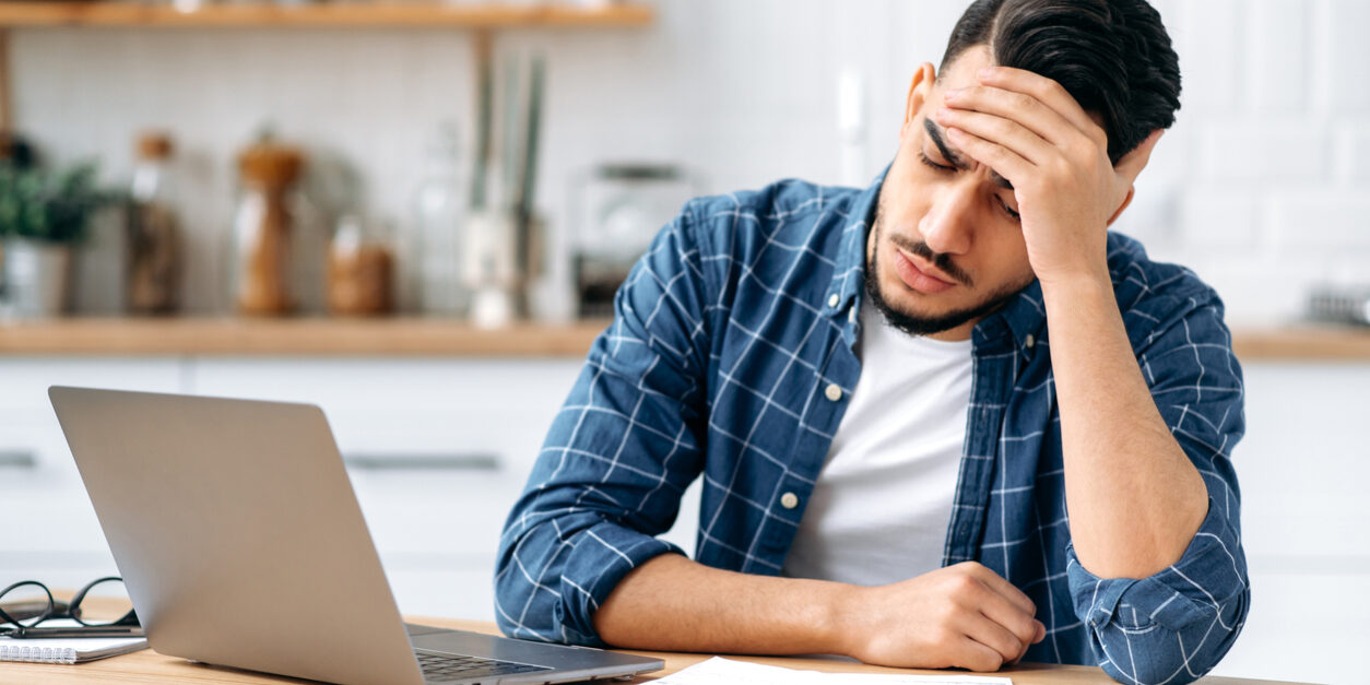 Tired worried sad arabian or indian guy, sit at a desk, looking tired and stressed, frustrated looking at work documents, failing to finish work on the project on time, needs rest. Overworking concept